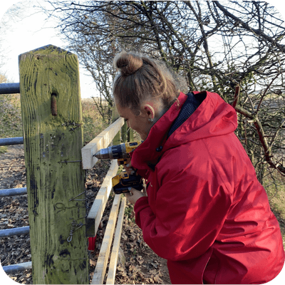 Woman in red jacket using a drill to fix a wooden piece on a gate outdoors, surrounded by trees.
